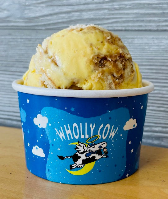 Wholly Cow Bananas Pudding flavor ice cream in a blue 'Wholly Cow' branded container on a wooden surface with a gray background.