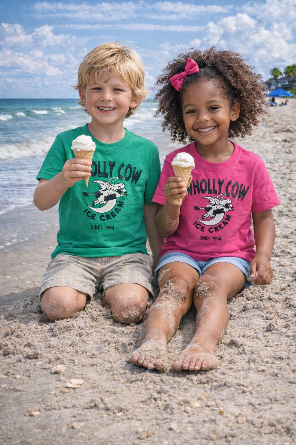 Two children sitting on Edisto beach eating local Charleston made wholly cow ice cream cones, wearing 'Wholly Cow Ice Cream' shirts.