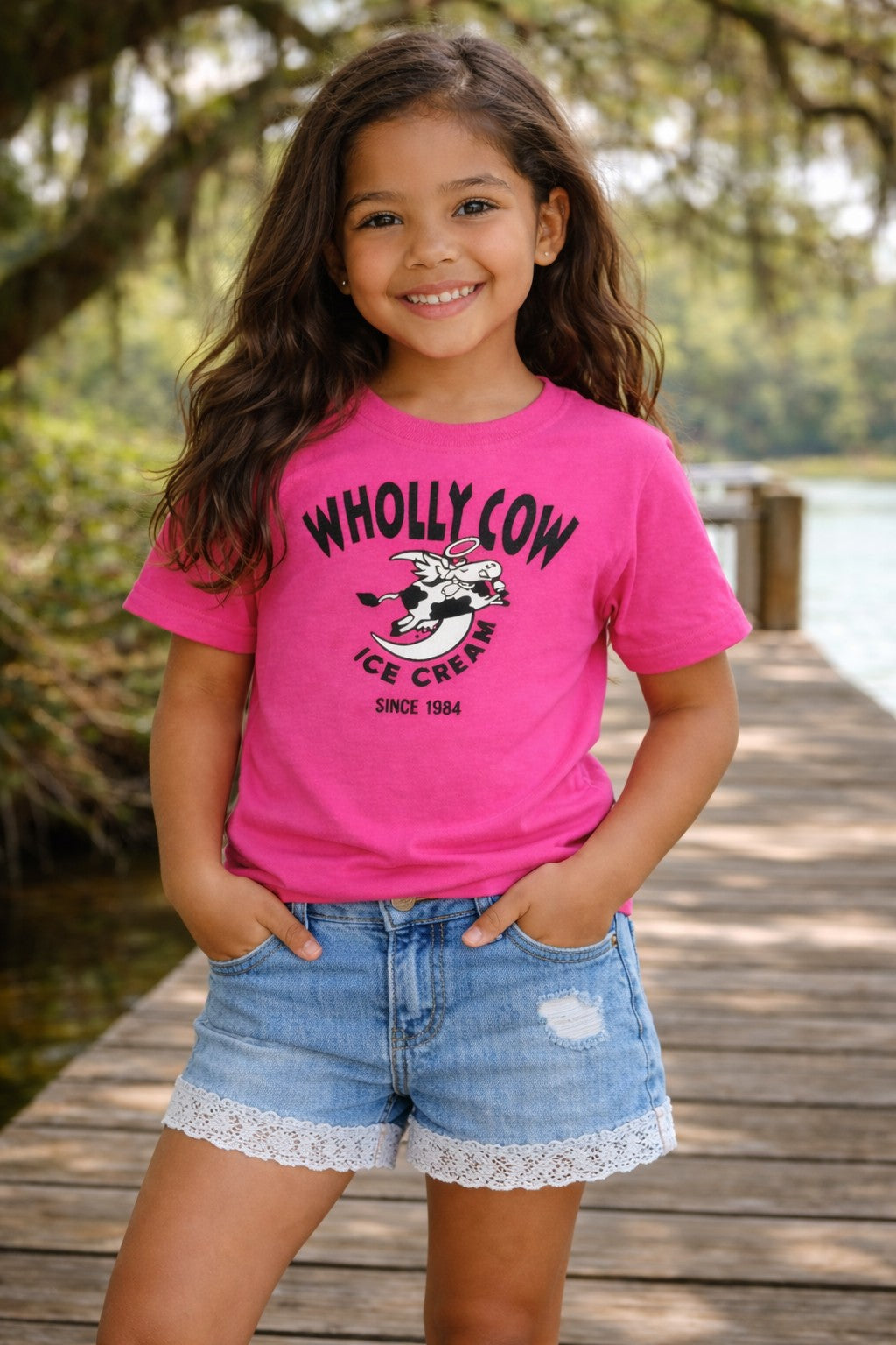 Young girl wearing a pink 'WHOLLY COW ICE CREAM' t-shirt and denim shorts on a wooden dock.