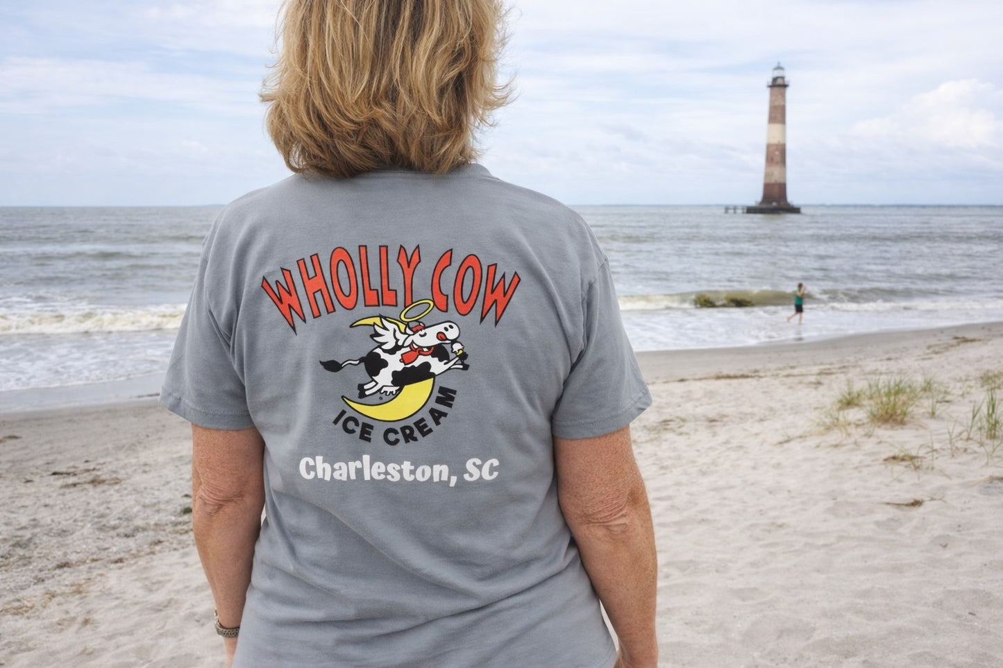 woman wearing a 'Wholly Cow Ice Cream Charleston, SC' t-shirt on a beach with a lighthouse in the background.