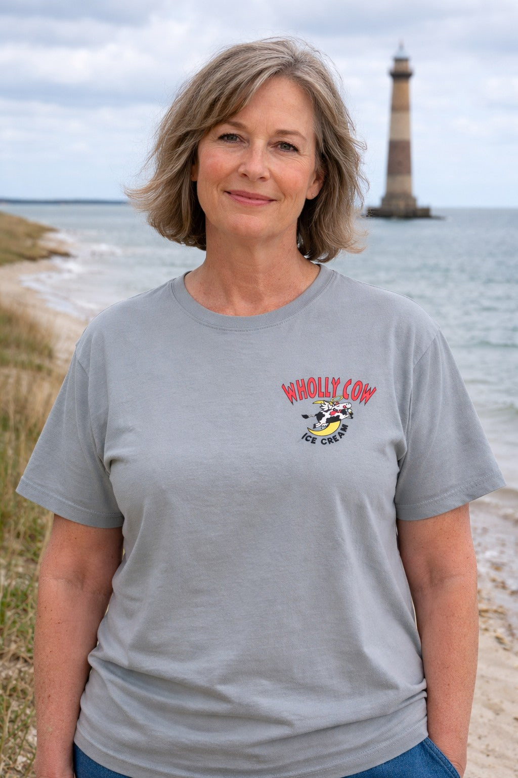Woman wearing a gray t-shirt with wholly cow logo on a beach with a lighthouse in the background